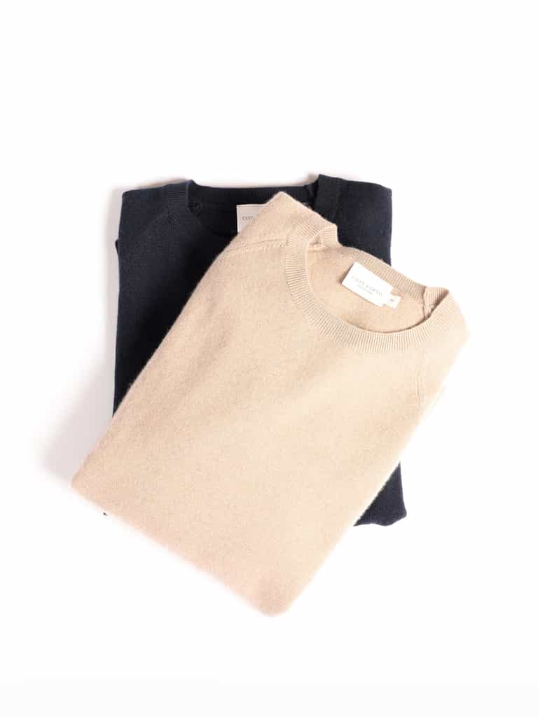 Two neatly folded Cozy Earth Men's Crewneck Sweaters placed on a white background. The top sweater is light beige, and the one underneath is dark navy. Each sweater has a small white label near the collar with text on it. |Color:Sandstone