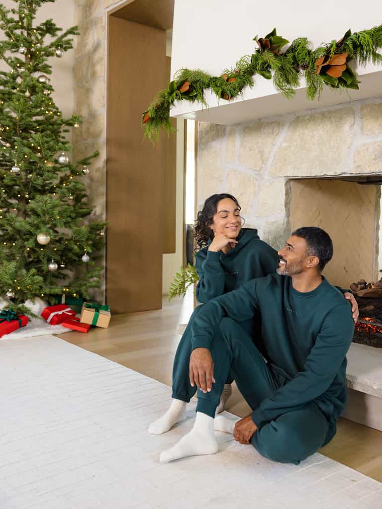 Dressed in matching Men's CityScape Crewnecks by Cozy Earth, a couple sits by the fireplace with warm smiles. A beautifully decorated Christmas tree and an array of gifts are visible in the background, while the mantel boasts lush greenery, creating a cozy and festive holiday atmosphere. |Color:Spruce