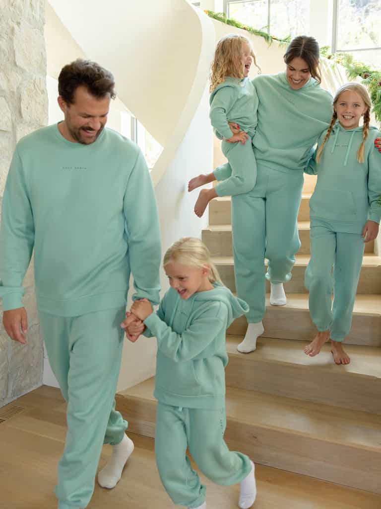 A family of five, all wearing matching Men's CityScape Crewnecks from Cozy Earth, cheerfully descends a wooden staircase. One adult carries a young child, while the others walk hand in hand. The space is filled with bright, natural light. |Color:Wintergreen