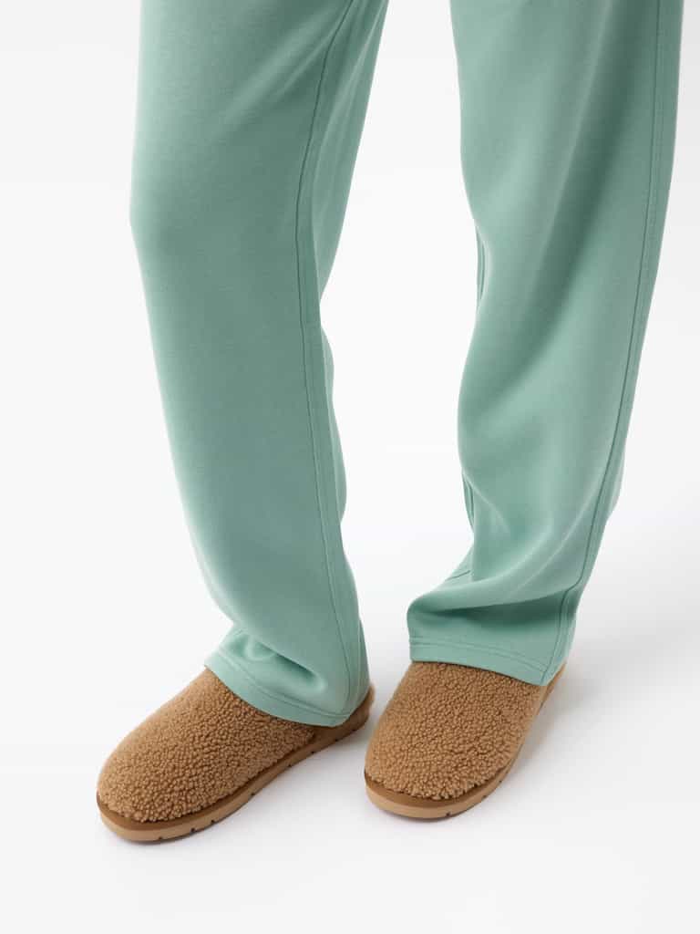A person dressed in Cozy Earth's light green Women's CityScape Wide Leg Pant and brown sherpa-lined slippers is standing against a white background. The pants fully cover the person's legs, while the slippers have a soft, fuzzy look. |Color:Wintergreen