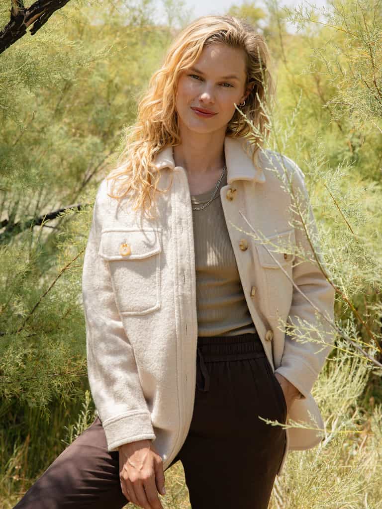 A blonde woman stands outdoors in front of greenery, wearing a light-colored Women's Boucle Shacket by Cozy Earth over a beige top and dark pants. Her wavy hair cascades over her shoulders, and she smiles gently while resting one hand against the side of her shacket. |Color:Alabaster