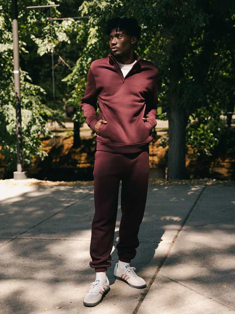 A person stands outdoors on a sunlit sidewalk, wearing Cozy Earth's Men's CityScape Quarter Zip in maroon and white sneakers. They have their hands in their pockets and are looking off to the side. The background features trees with green foliage, casting dappled shadows on the ground. |Color:Burgundy