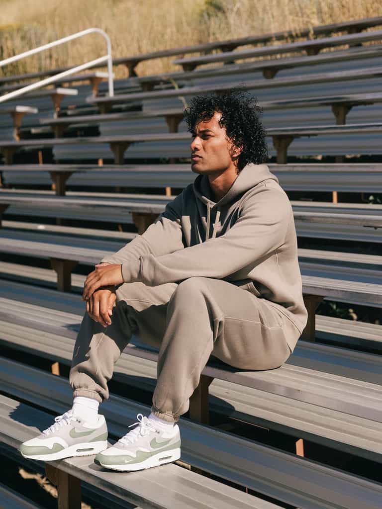 A person with curly hair sits on an empty set of bleachers outdoors, wearing a light gray hoodie and matching Cozy Earth Men's CityScape Sweatpants, paired with green and white Nike sneakers. They look off into the distance with a thoughtful expression. |Color:Ebony