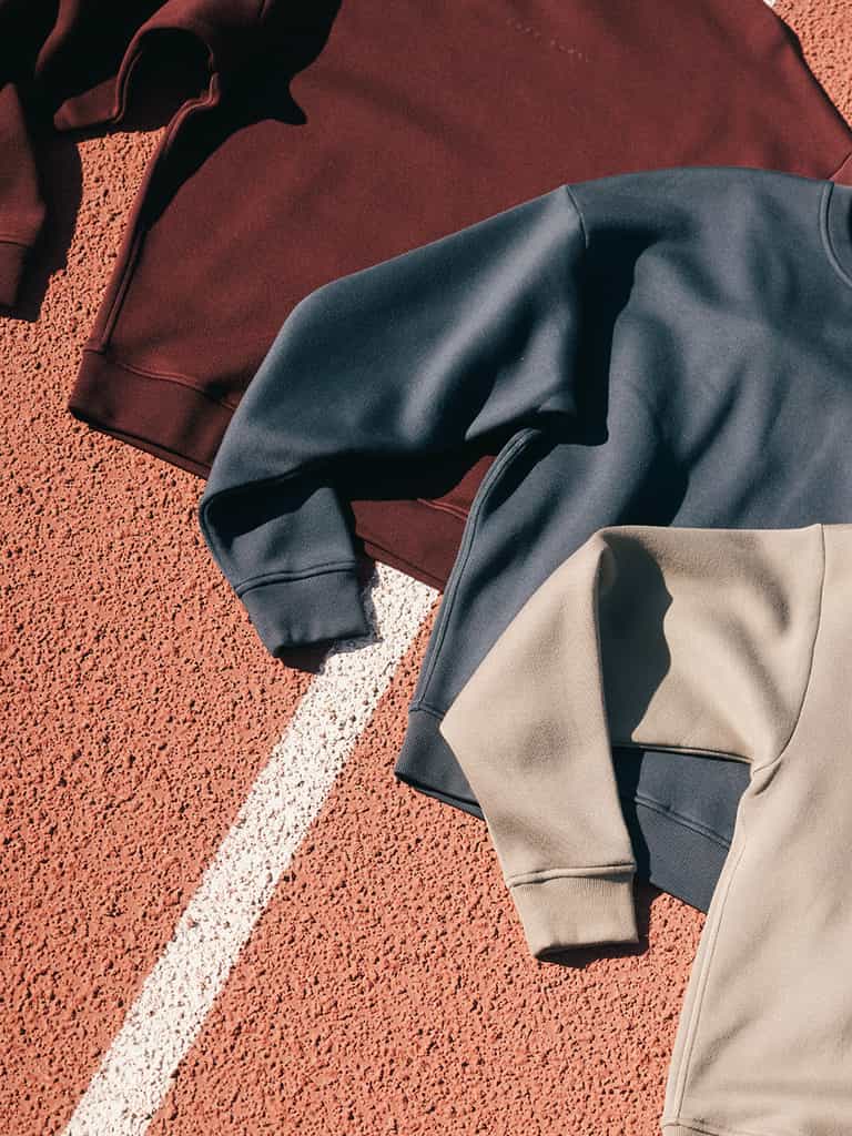 Various Women's CityScape Crewneck sweatshirts by Cozy Earth are laid out on a textured surface resembling a running track. The sweatshirts include dark red, dark gray, and beige tones, with sleeves slightly overlapping each other on the lined surface. |Color:Eclipse