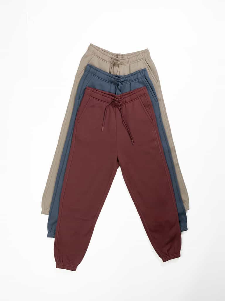 Three pairs of Men's CityScape Sweatpants by Cozy Earth are displayed in an overlapping arrangement. The sweatpants on top are maroon, the middle pair is dark blue, and the bottom pair is light beige. Each pair features a drawstring waistband and elastic cuffs. The background is white. |Color:Eclipse