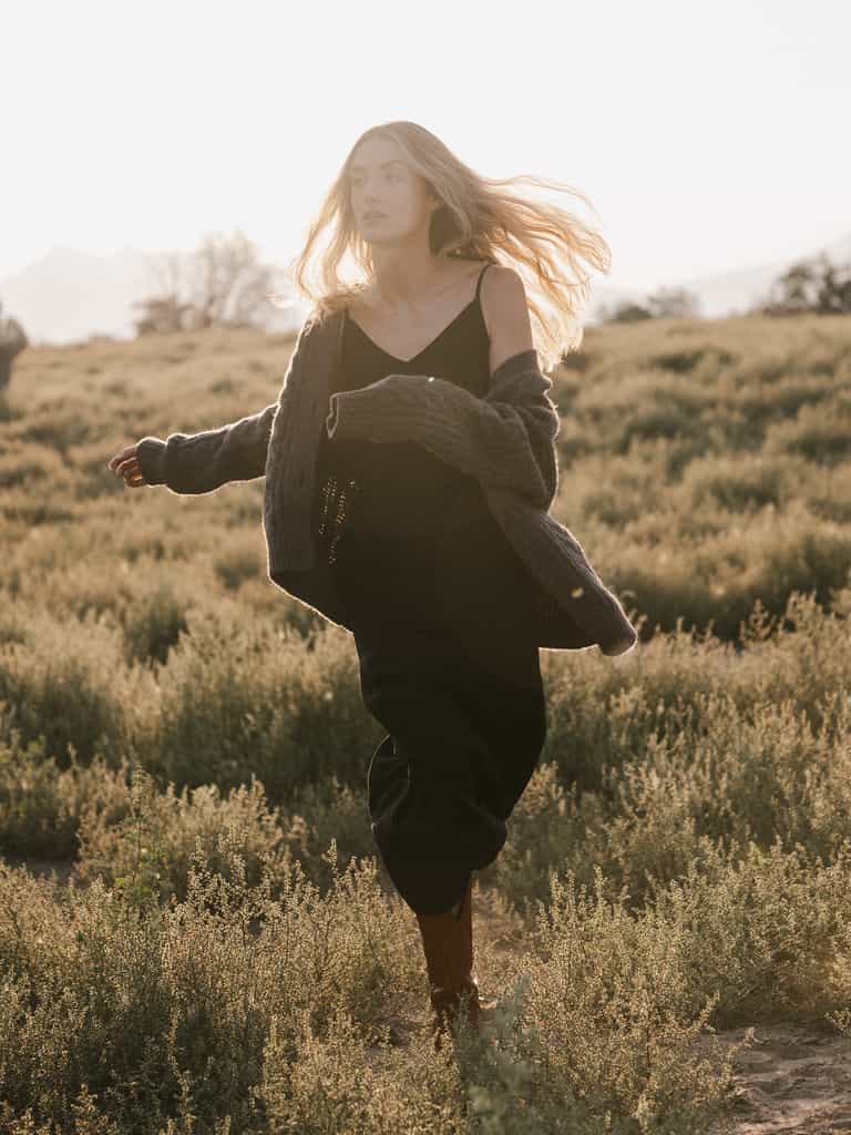 A woman with long hair walks through a grassy field during sunset. She is wearing the Women's Sunset Slip Dress from Cozy Earth, paired with a large, cozy sweater that is partially off her shoulders. Her hair flows in the breeze as she gazes forward, creating a serene and peaceful scene. |Color:Eclipse