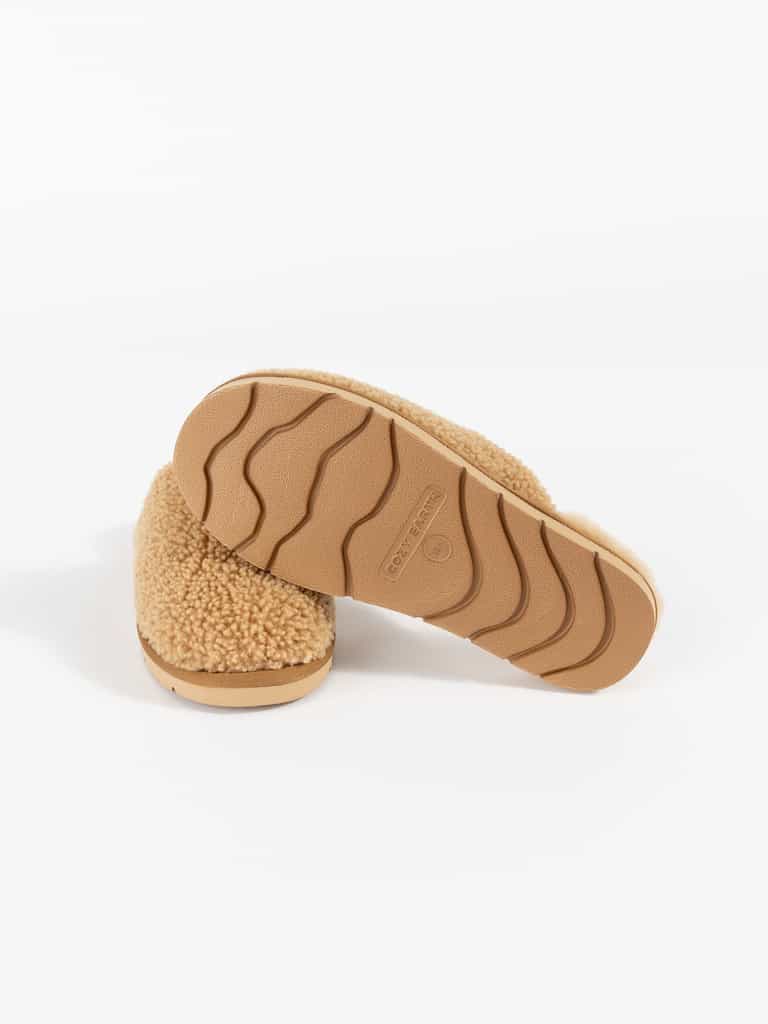 A pair of Cozy Earth's Puffy Sheep Slippers, featuring a tan fleece texture and embossed rubber soles with a wavy grip pattern, displayed in a studio setting. The slippers are arranged to showcase both the top and bottom views. |Color:Fawn