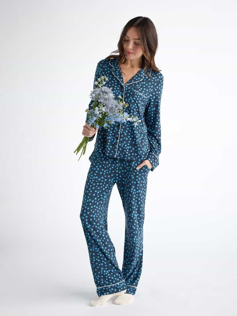 A person in Cozy Earth Women's Bamboo Stretch-Knit Pajama Pant with blue polka dots and white piping stands barefoot against a white background, holding a bouquet of flowers. |Color:Forget Me Nots