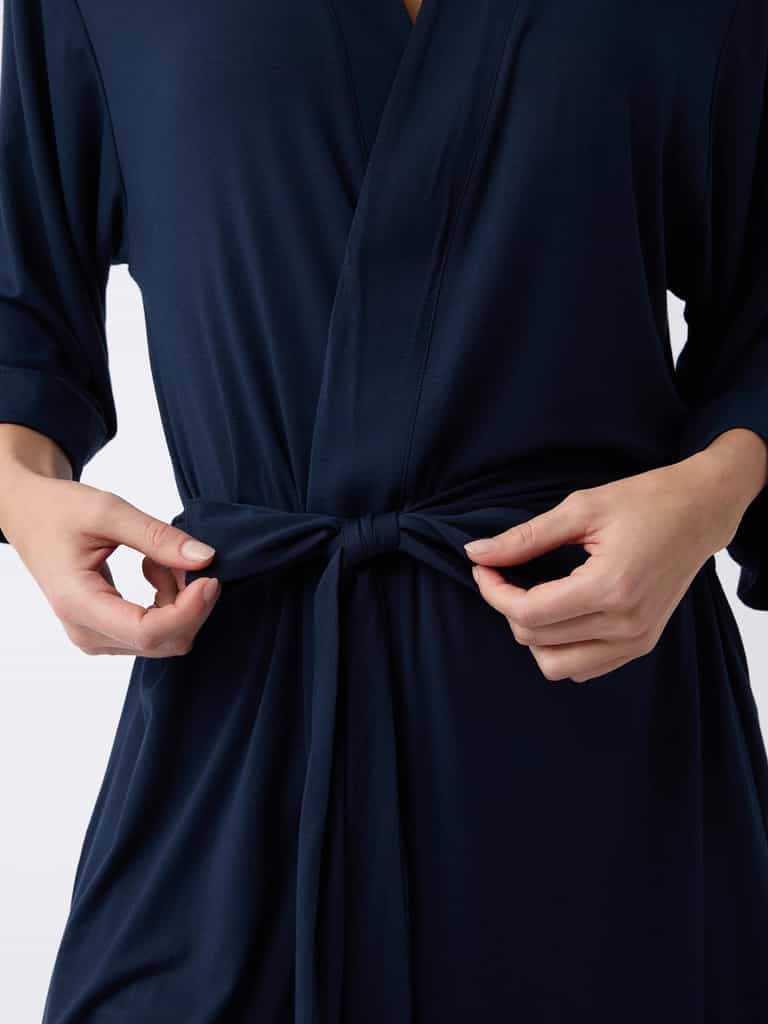 An individual secures the belt of a Cozy Earth Women's Stretch-Knit Bamboo Kimono Robe in dark blue, highlighting their hands and the soft, long-sleeved fabric. |Color:Navy