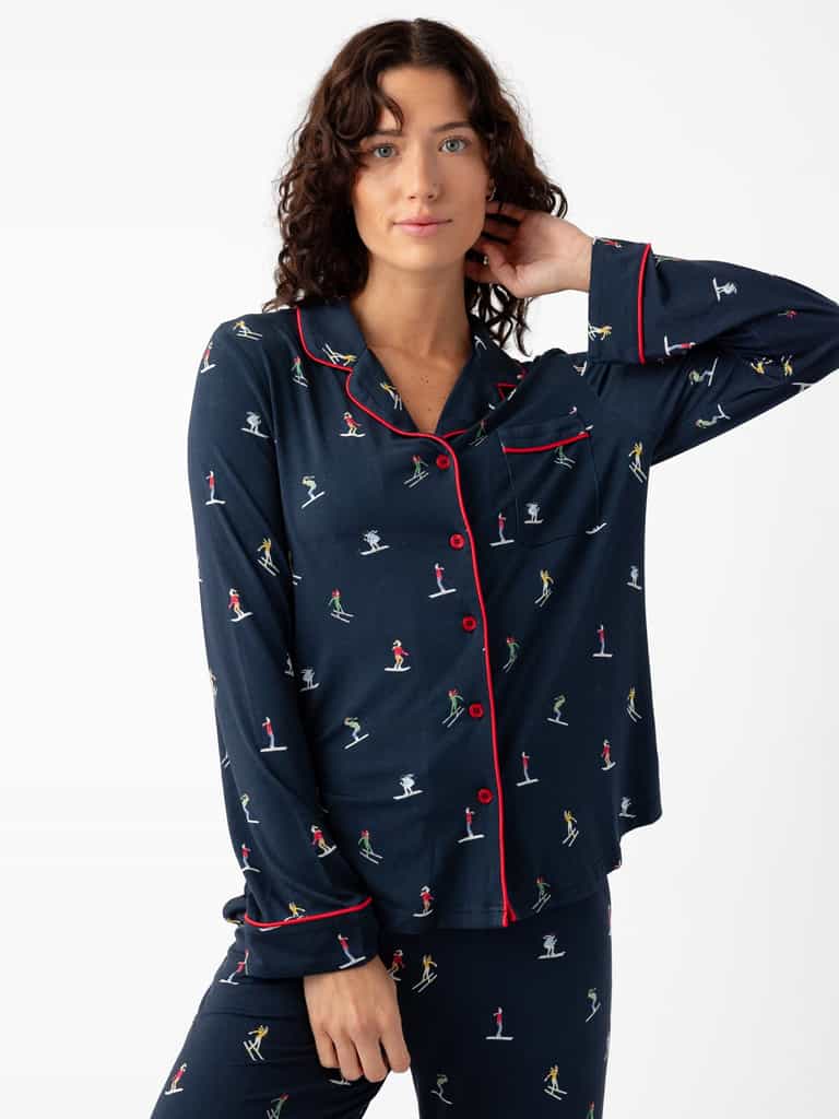 A person in the Cozy Earth "Last Chance" Women's Bamboo Stretch-Knit Long Sleeve Pajama Set, featuring a navy blue color with red buttons and multicolored prints of people engaged in activities, poses against a plain white background with curly hair. |Color:Skiers Navy