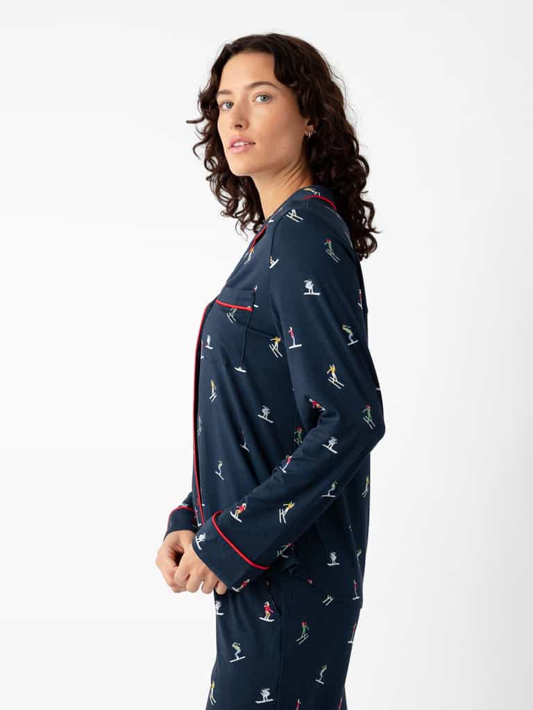 A person with long curly hair stands sideways, wearing the Cozy Earth Last Chance Women's Bamboo Stretch-Knit Long Sleeve Pajama Set in navy with colorful skier patterns and red piping, looking over their shoulder against a plain white background. |Color:Skiers Navy