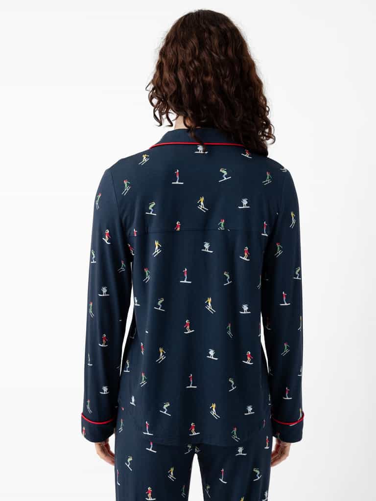 A person with long, curly hair stands facing away, wearing Cozy Earth's Last Chance Women's Bamboo Stretch-Knit Long Sleeve Pajama Set featuring small colorful figures and red piping on the collar and cuffs. The set comes with matching bottoms, all set against a plain white background. |Color:Skiers Navy