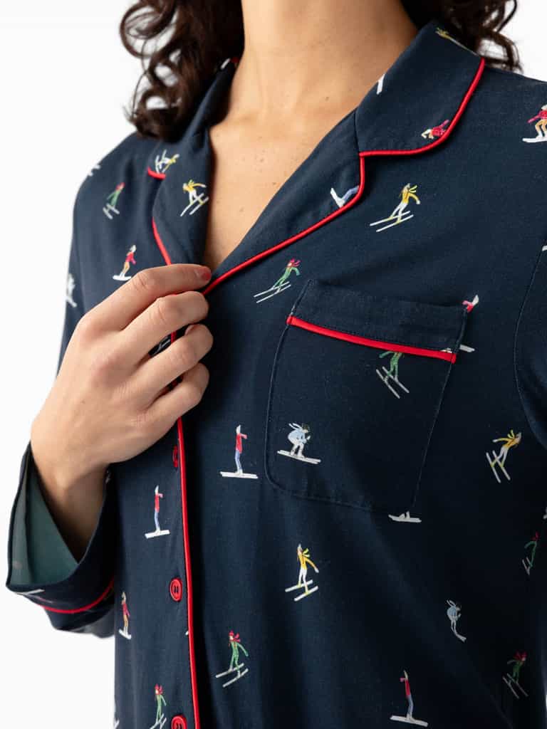 A person adjusts the collar of a Cozy Earth's Last Chance Women's Bamboo Stretch-Knit Long Sleeve Pajama Top, featuring a skiing print with red piping and buttons. |Color:Skiers Navy