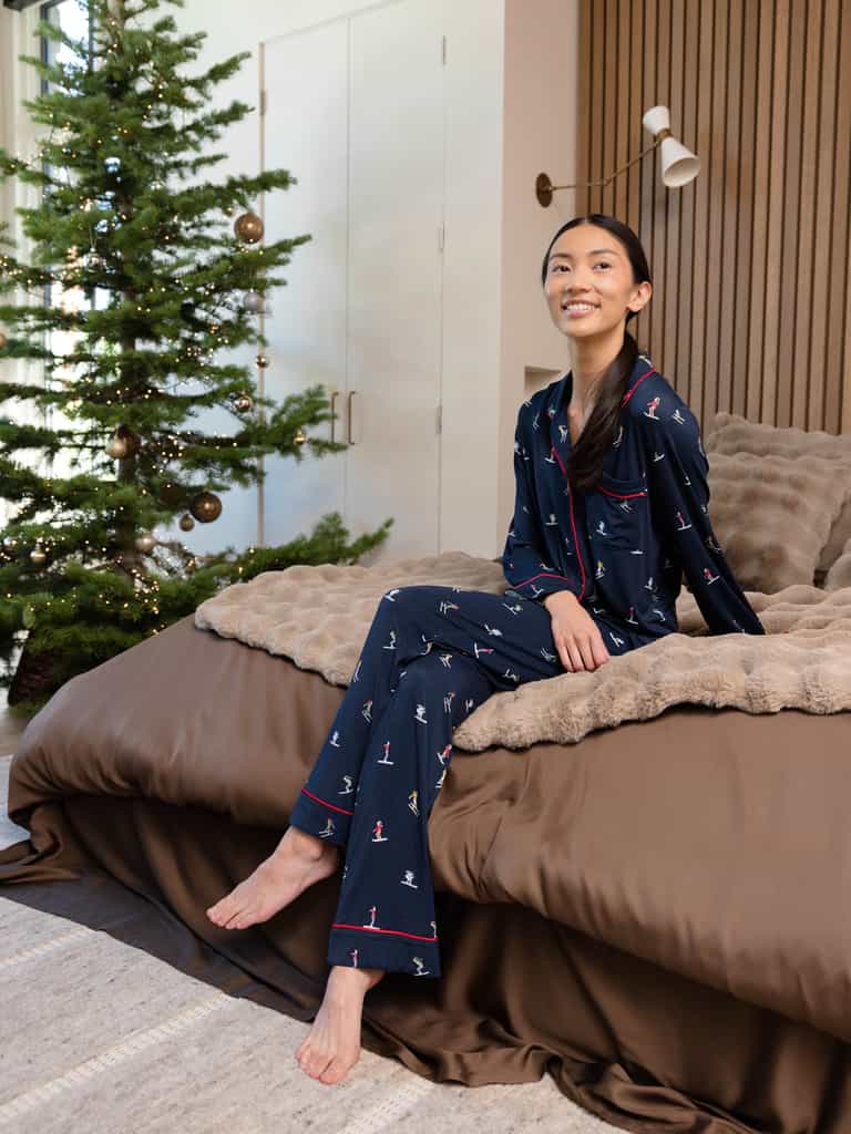 A person wearing Cozy Earth's Last Chance Women's Bamboo Stretch-Knit Long Sleeve Pajama Set in dark blue sits smiling on the bed's edge. The cozy room features a fluffy blanket and a decorated Christmas tree in the background. |Color:Skiers Navy