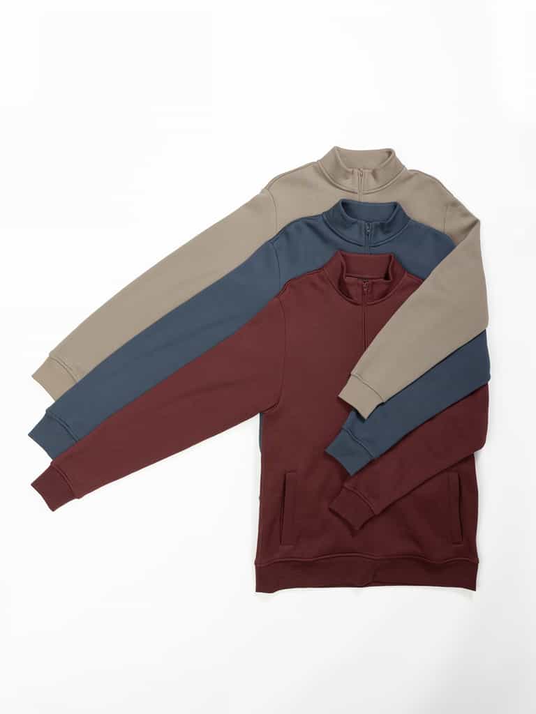 Three Men's CityScape Quarter Zip sweaters from Cozy Earth are laid on a white surface. The top sweater is beige with dark gray sleeves, the middle one is gray with teal sleeves, and the bottom one is maroon. They have a casual, comfortable appearance. |Color:Burgundy