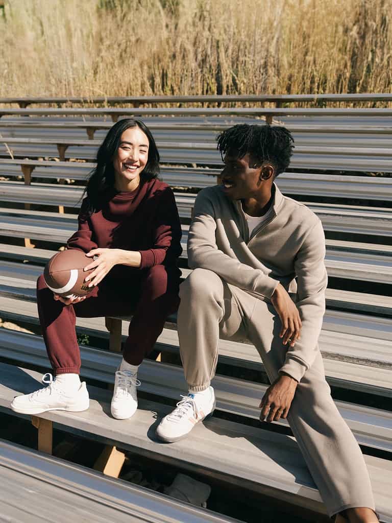 Two people sit on bleachers outside on a sunny day. The person on the left, wearing a red CityScape Quarter Zip by Cozy Earth, smiles while holding an American football. The person on the right, dressed in a beige CityScape Quarter Zip by Cozy Earth, smiles and looks at the other person. Both wear white sneakers. |Color:Ebony