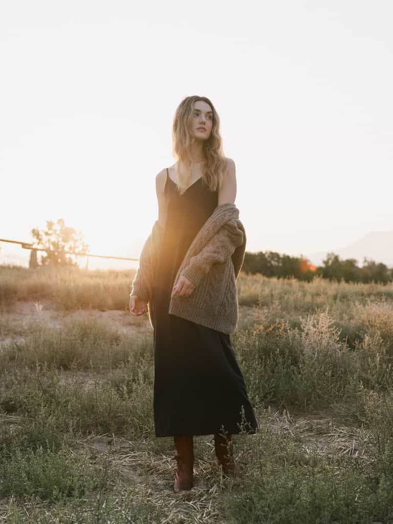 A woman with long wavy hair stands in a field at sunset. She is wearing Cozy Earth's Women's Sunset Slip Dress, a cozy brown cardigan draped over her shoulders, and brown boots. She gazes off into the distance as the sun casts a warm glow around her. |Color:Eclipse