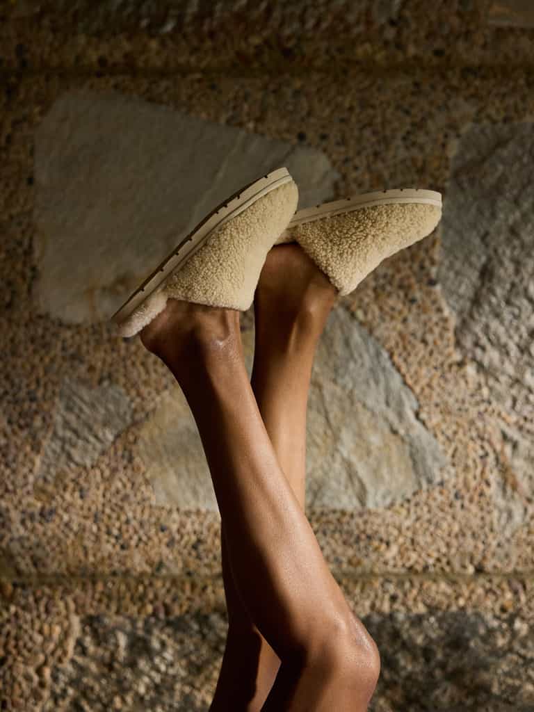 A person crosses their legs upwards, showcasing beige Puffy Sheep Slippers by Cozy Earth against a textured stone wall backdrop. The slippers have a cozy, soft appearance with sturdy soles. The ambient lighting casts gentle shadows, emphasizing the texture and warmth of the scene |Color:Natural.