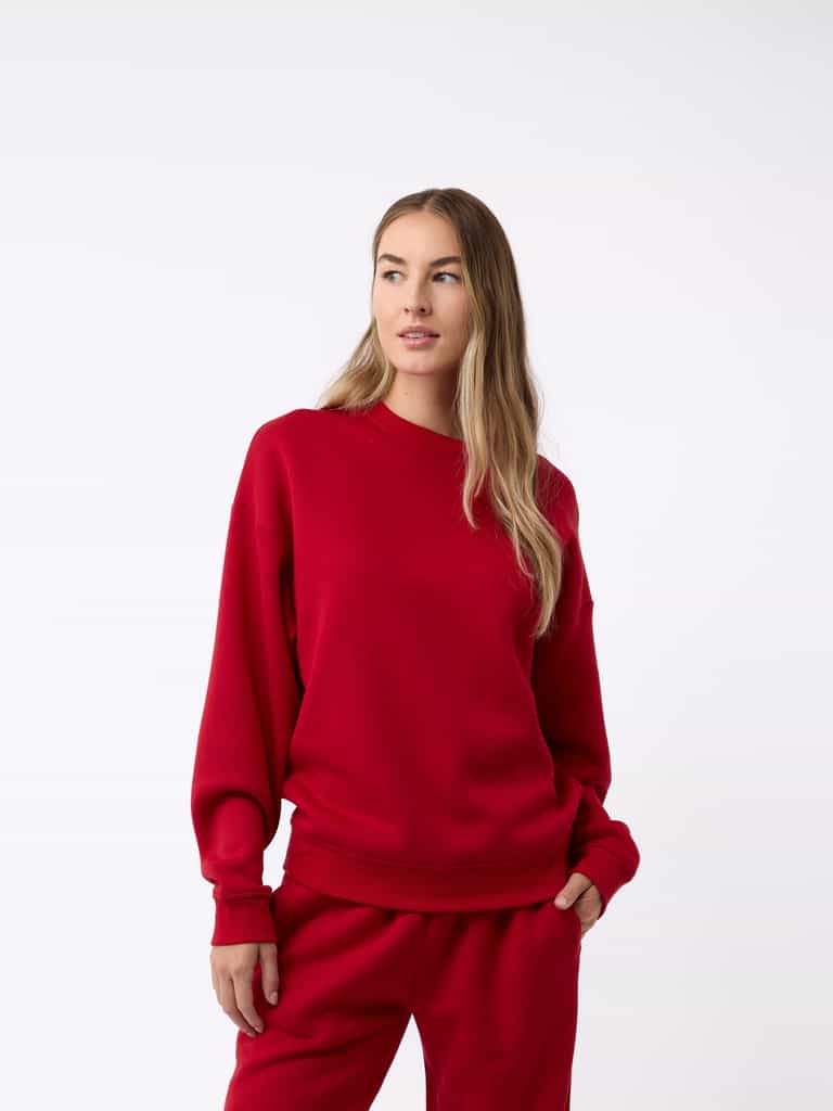 A person with long hair is wearing a Cozy Earth Women's CityScape Crewneck and matching sweatpants, standing against a plain white background. They have one hand in their pocket and are gazing slightly to the side. |Color:Crimson