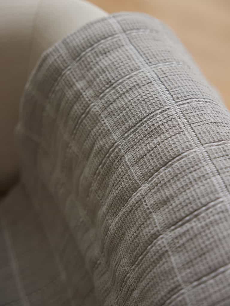 A close-up image of Cozy Earth's beige-colored Waffle Windowpane Blanket draped over part of a light-colored upholstered chair. The blanket's grid-like, waffle pattern creates a soft and cozy appearance. The background is softly blurred. |Color:Dove Grey
