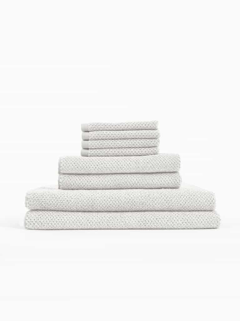 The Cozy Earth Nantucket Bath Towel Set features textured, light gray towels in assorted sizes, neatly stacked on a white background.|Color:Heathered Light Grey