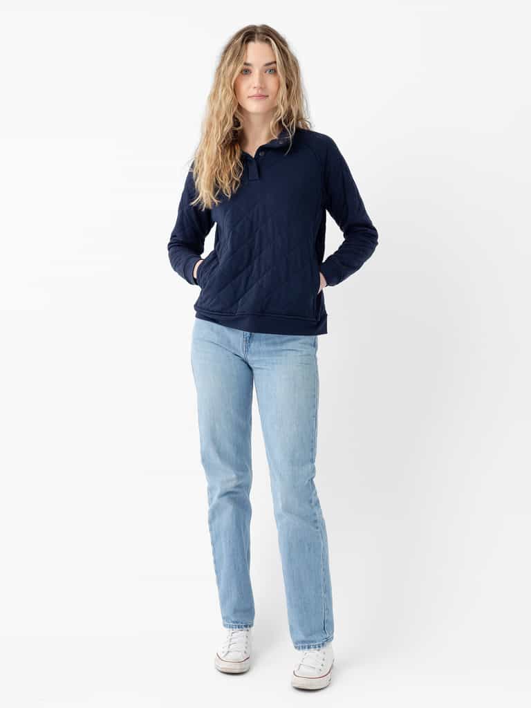 A person with long, wavy hair is wearing a Cozy Earth Women's Ultra-Soft Bamboo Quilted Snap Pullover in navy blue, light blue jeans, and white sneakers. They are standing against a plain white background with their hands in their pullover pockets, looking directly at the camera. |Color:Navy