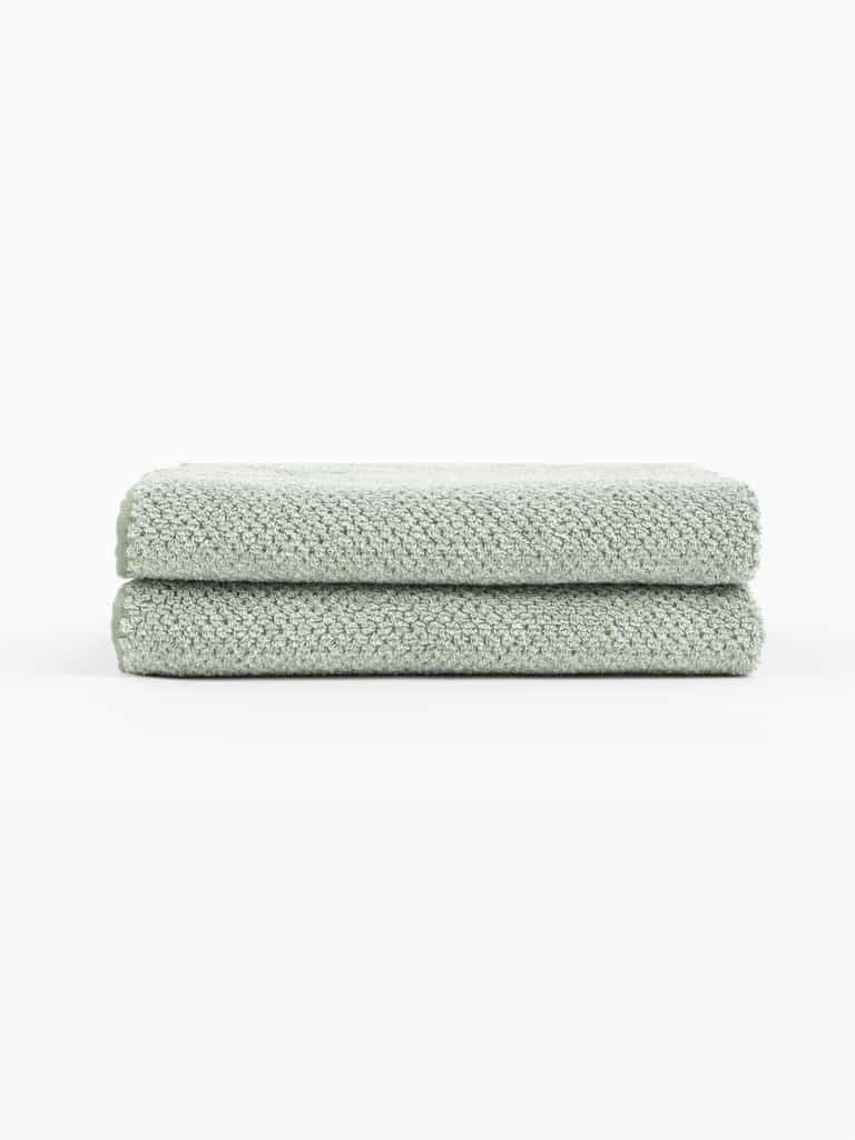 Two Nantucket Bath Towels by Cozy Earth, in a light green textured finish, are neatly folded and stacked on a white surface with a plain white background.|Color:Heathered Sage