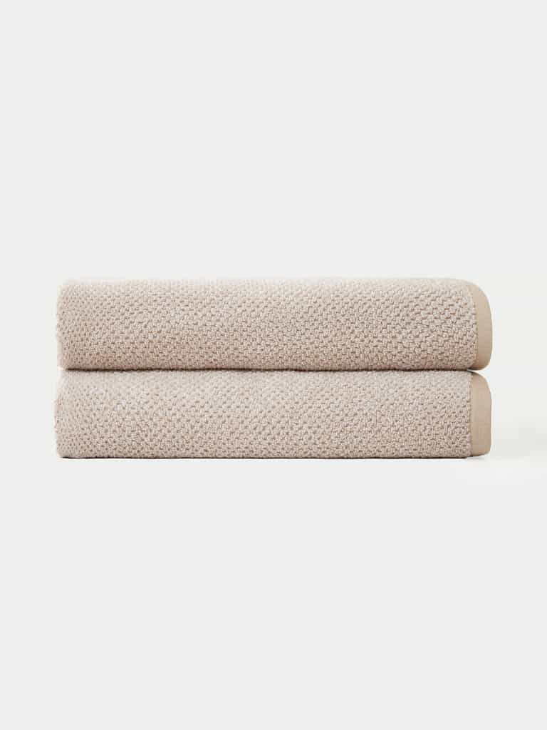 Two Nantucket Bath Towels by Cozy Earth are neatly folded and stacked together against a plain white background.|Color:Heathered Sand