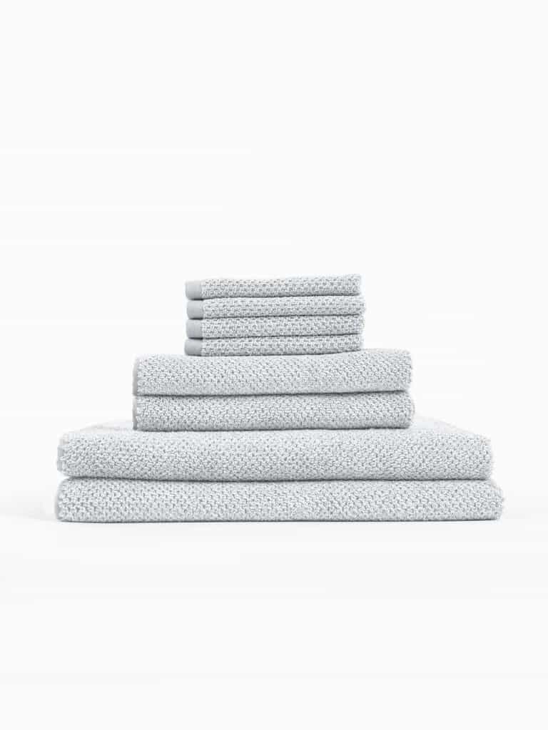 The Cozy Earth Nantucket Bath Towel Set features light grey textured towels in multiple sizes, neatly stacked from largest to smallest against a plain white background.|Color:Heathered Harbor Mist