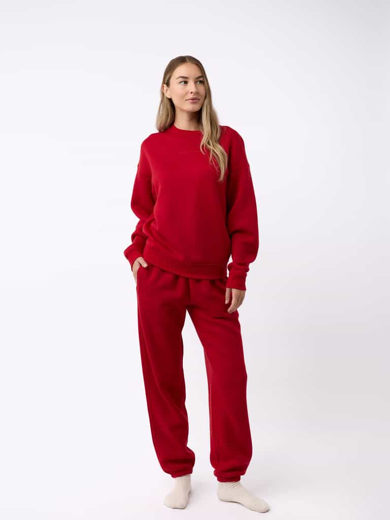 A person wearing Cozy Earth's Women's CityScape Crewneck and matching red sweatpants stands against a plain white background. With long hair, they are looking slightly to the side and have light-colored socks on as well. |Color:Crimson