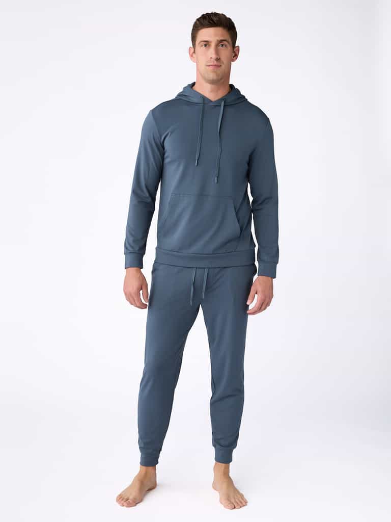 A man with short, dark hair and a neutral expression stands barefoot on a white background, dressed in Cozy Earth's Men's Ultra-Soft Bamboo Pullover Crew in teal. The pullover features a front pocket and drawstrings, while the matching jogger pants are fitted and comfortable. |Color:Dawn