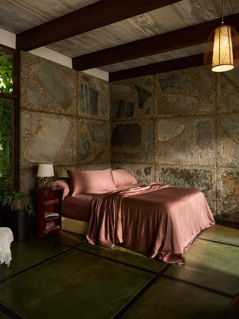 A cozy, rustic bedroom featuring a bed with silky, Bamboo Flat Sheets by Cozy Earth in rose color against a textured stone wall. Wooden beams line the ceiling, while a woven pendant light hangs above. Nearby, there is a red nightstand with a lamp, and a lush plant adds a touch of greenery. |Color:Terra