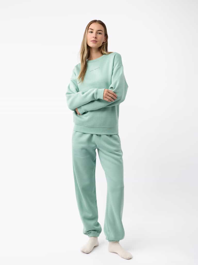 A woman wearing the Women's CityScape Crewneck by Cozy Earth stands gracefully in a light green sweatshirt and sweatpants. With long flowing hair and a direct gaze fixed on the camera, she embodies a casual and comfortable style, standing barefoot against a plain white backdrop. |Color:Wintergreen