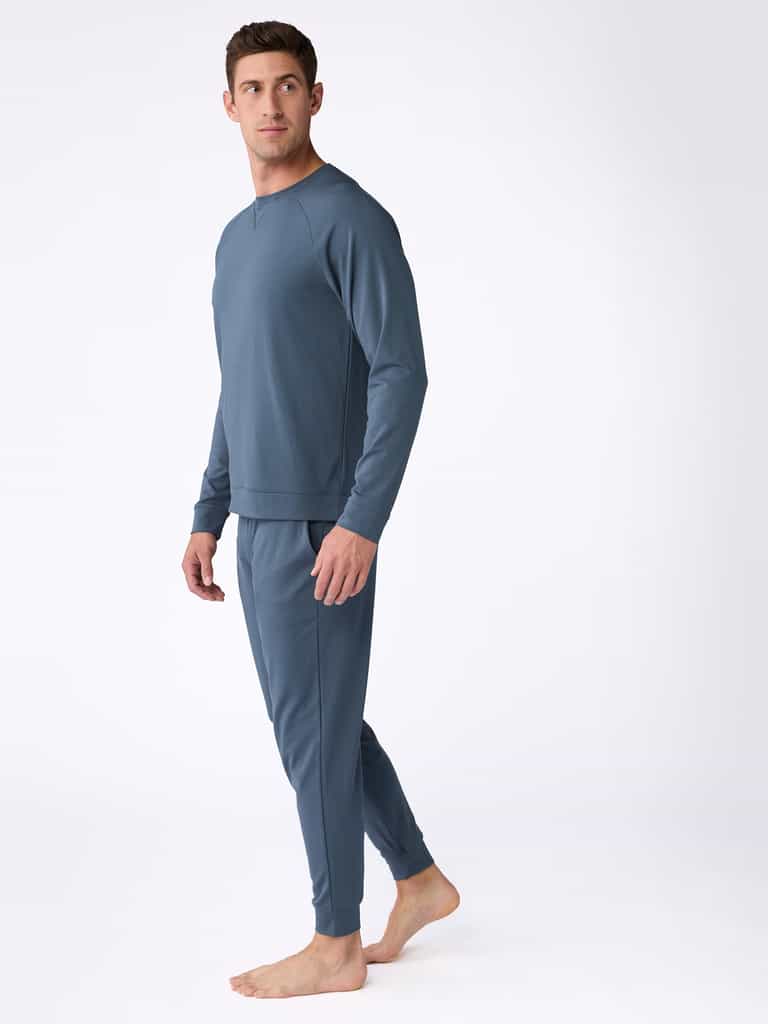 A person is barefoot on a white background, wearing the Men's Ultra-Soft Bamboo Pullover Crew by Cozy Earth in a matching blue set that includes long-sleeve tops and pants. The individual gazes to the side, one foot slightly forward, showcasing the outfit's comfortable and casual style. |Color:Dawn