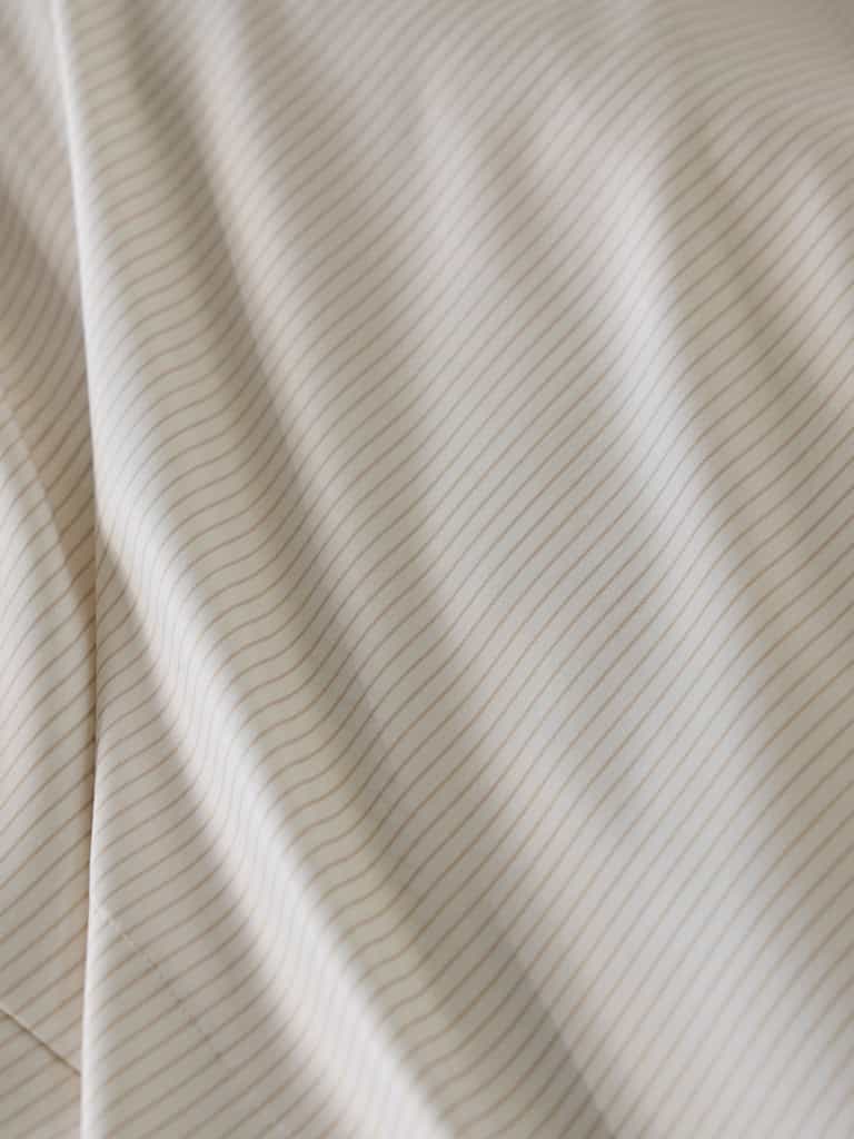 Close-up of the Bamboo Flat Sheet by Cozy Earth, featuring a beige fabric with thin, evenly spaced horizontal lines in a light brown color. The fabric appears to be soft and smooth, with gentle folds and subtle shadows enhancing its texture. |Color:Driftwood Pinstripe
