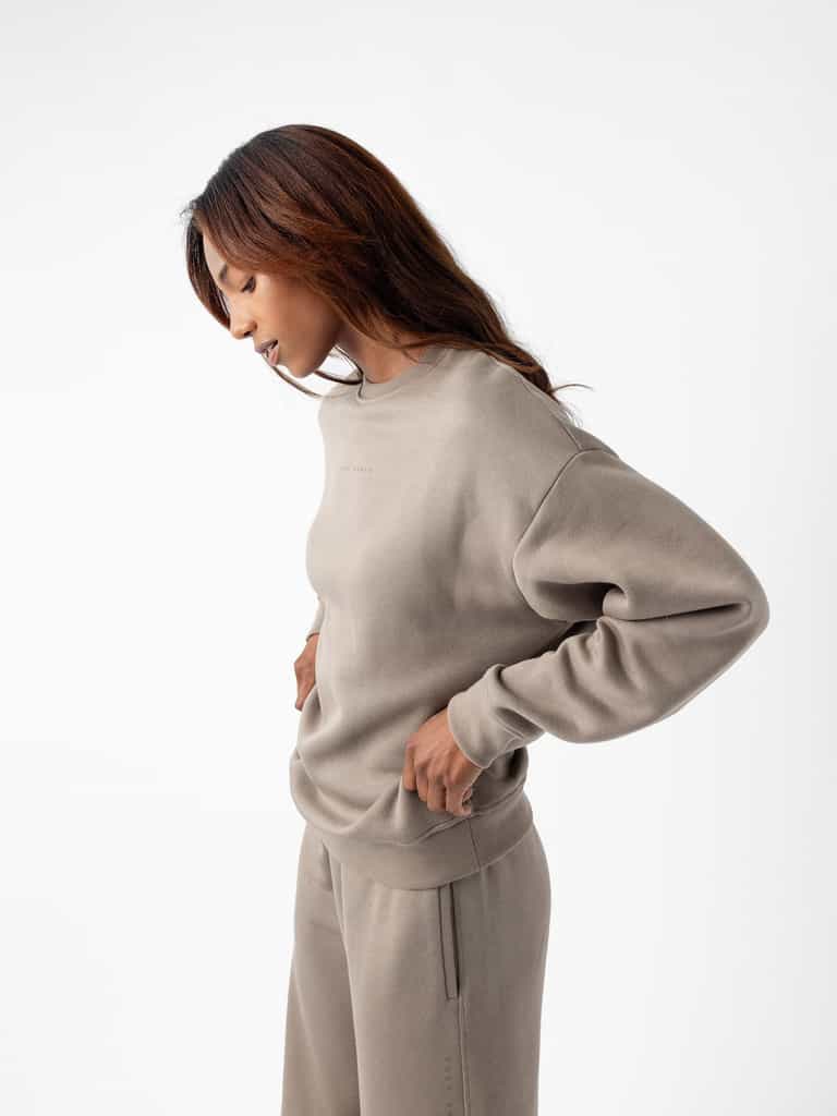 A woman with long, brown hair is standing in profile against a white background. She is wearing the Cozy Earth's Women's CityScape Crewneck sweatshirt and matching pants in light brown, with her hands resting on her hips and looking downward. The outfit appears comfortable and casual. |Color:Ebony