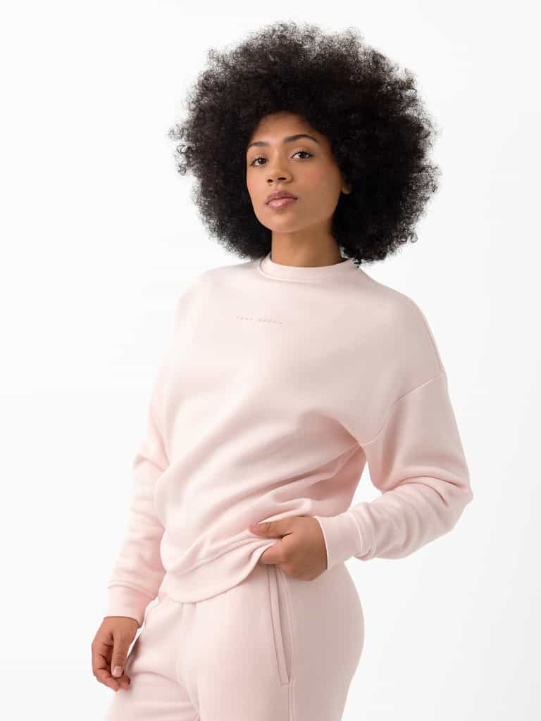 A person with curly hair stands against a white background, wearing Cozy Earth's Women's CityScape Crewneck and matching pants. They have one hand in their pocket and a relaxed expression. |Color:Peony