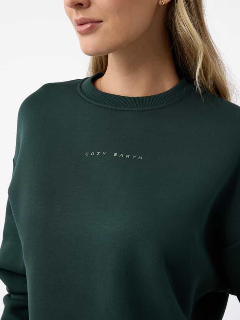 A person wearing a Women's CityScape Crewneck sweatshirt in dark green from Cozy Earth. The individual, with long, light-colored hair, is facing slightly to the side against a plain background. |Color:Spruce