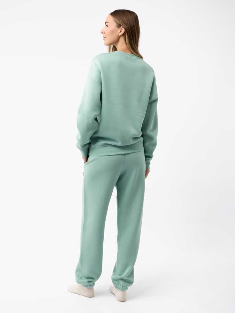 A person with long hair stands facing away, wearing the mint green Women's CityScape Crewneck and matching sweatpants from Cozy Earth. The outfit is casual and cozy, complemented by white sneakers, set against a plain white background. |Color:Wintergreen