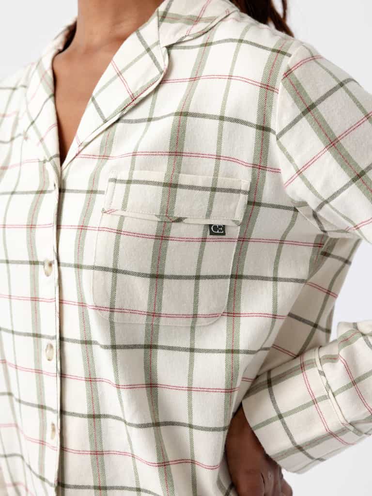 A person wearing a cream-colored plaid shirt from the Cozy Earth Women's Farmhouse Long Sleeve Pajama Set, featuring green and pink lines, stands with one hand on their hip. The shirt includes a button-up front and a chest pocket with a small tag. The background is plain white. |Color:Alabaster/Juniper