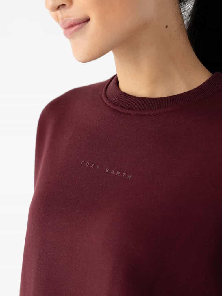 A person wearing a Women's CityScape Crewneck in deep red, with the brand name "Cozy Earth" subtly printed on the chest. The image focuses on the upper torso and part of the face against a plain white background. |Color:Burgundy