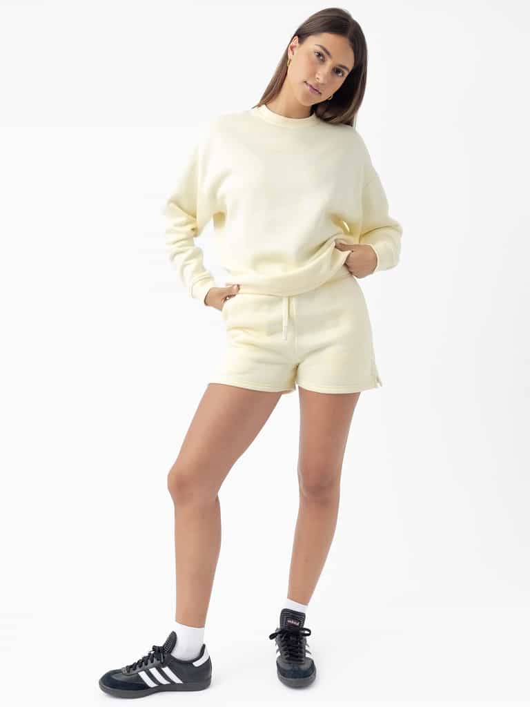 A woman with long hair stands in a relaxed pose wearing Cozy Earth's Women's CityScape Crewneck in light yellow along with matching shorts. She finishes the look with black sneakers and white socks against a plain white background. |Color:Lemonade