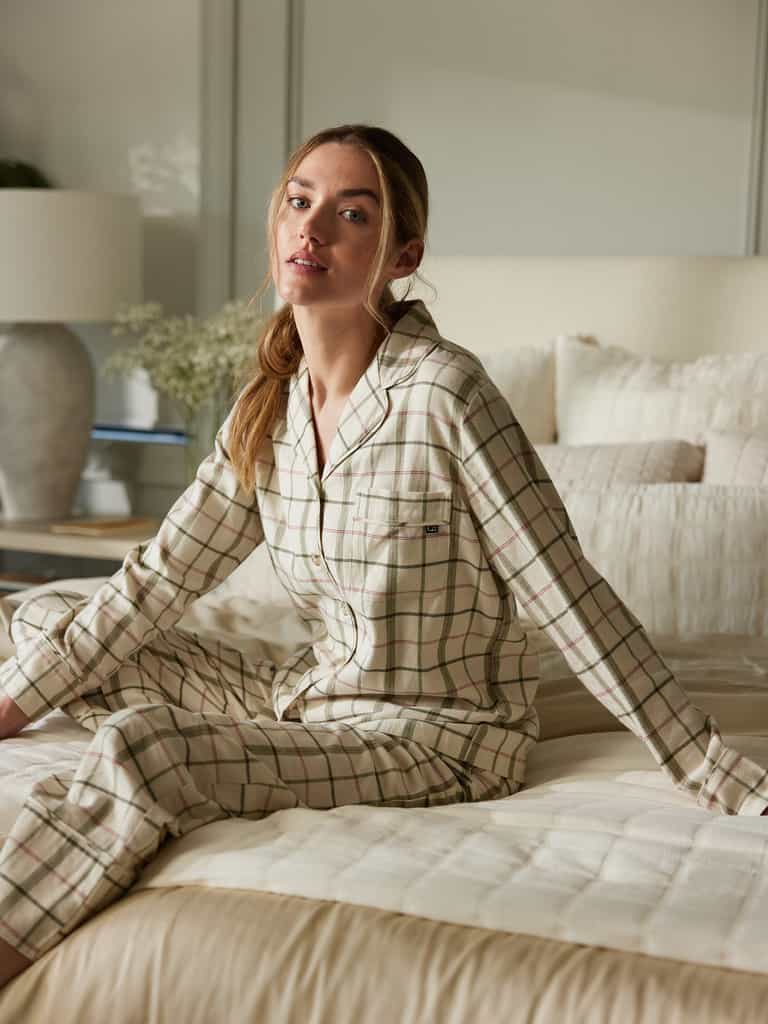 A woman with brown hair tied back sits on a bed in a cozy bedroom. She is wearing the Women's Farmhouse Long Sleeve Pajama Set from Cozy Earth and looking towards the camera. The bed is covered with a quilt, and a large lamp and decorative plant are visible in the background. |Color:Alabaster/Juniper