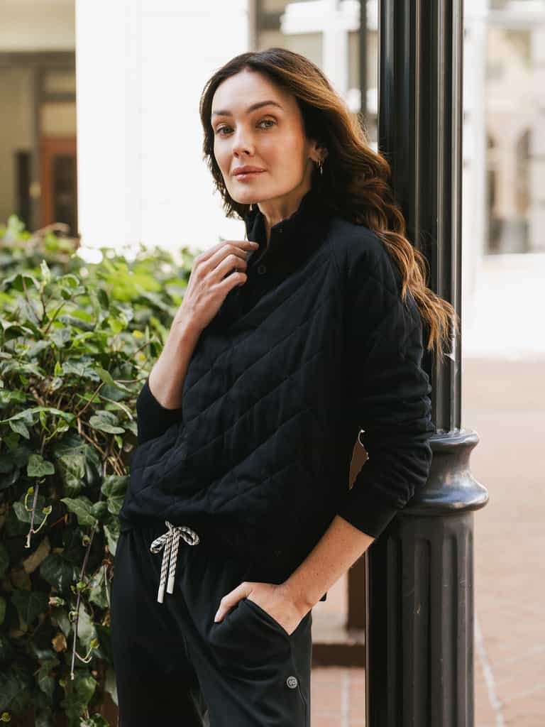 A woman with long, wavy hair leans against a lamppost outdoors. She is wearing Cozy Earth's Women's Ultra-Soft Bamboo Quilted Snap Pullover in black and black drawstring pants. Her right hand is near her neck, and her left hand is in her pocket. There are green plants and buildings in the background. |Color:Black