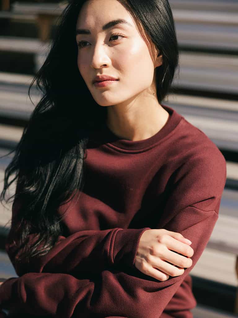 A person with long, dark hair, wearing a maroon Women's CityScape Crewneck by Cozy Earth, is sitting outdoors. Their arms are crossed as they gaze slightly to the side with a calm, contemplative expression. Sunlight illuminates their face, accentuating their features. |Color:Burgundy