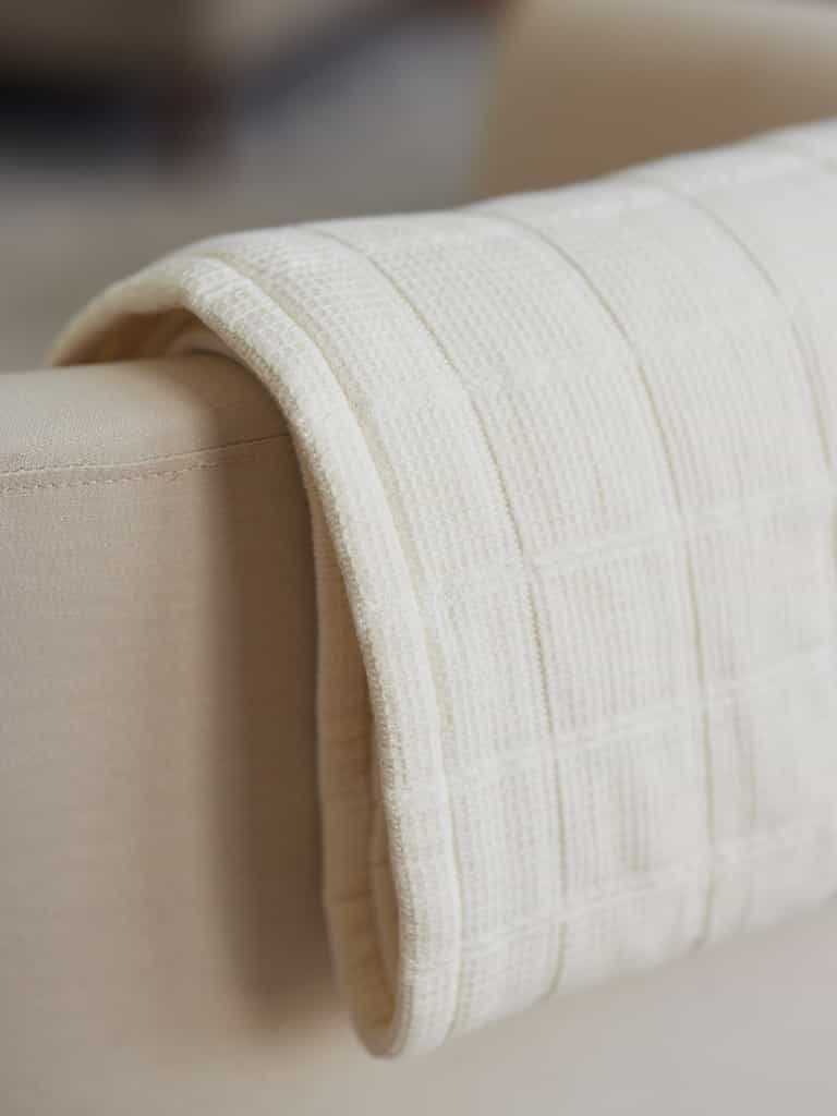 A *cream-colored, Waffle Windowpane Blanket* from Cozy Earth is delicately draped over the armrest of a beige sofa. This knitted throw blanket features a textured grid pattern, enhancing the cozy and warm aesthetic of the furniture piece. The background is softly blurred, keeping the focus on the blanket and sofa. |Color:Creme