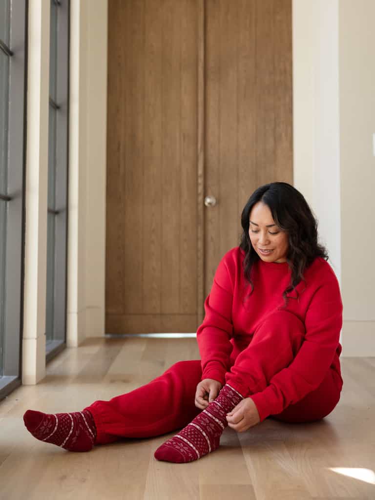 Dressed warmly in a red Women's CityScape Crewneck by Cozy Earth, an individual sits on a light wooden floor, adjusting their red patterned socks. Large wooden doors and tall windows in the background allow natural light to stream in. |Color:Crimson