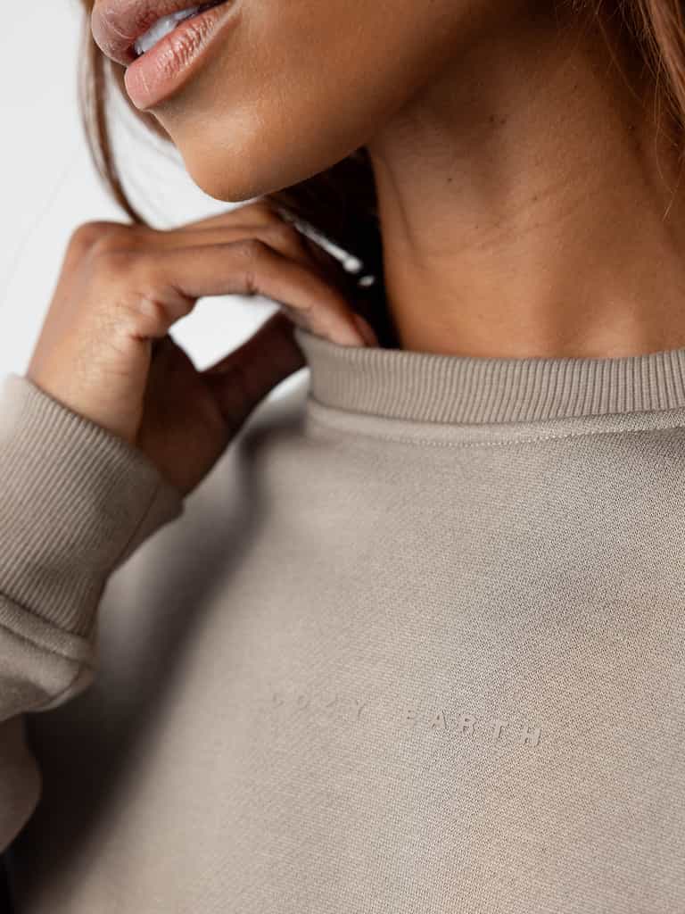 Close-up of a person wearing the Women's CityScape Crewneck in light grey, with "Cozy Earth" subtly embroidered across the chest. The person's hand is gently touching the neckline, and part of their face and neck is visible. |Color:Ebony