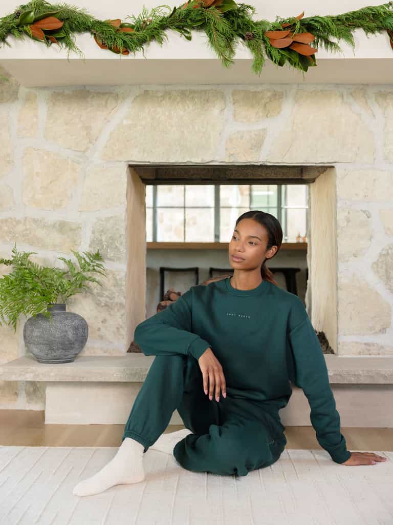 In a cozy stone-walled room, a woman sits on the floor wearing the Women's CityScape Crewneck by Cozy Earth in dark green, paired with matching sweatpants. A potted plant is beside her and festive greenery adorns the fireplace above. She appears relaxed and thoughtful. |Color:Spruce