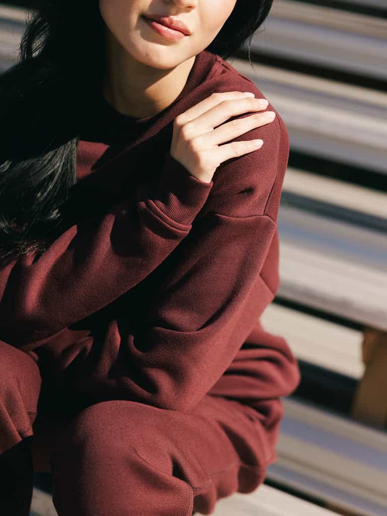 Wearing a Cozy Earth Women's CityScape Crewneck, a person is seated outdoors on wooden steps. They are touching their left shoulder with their right hand, and only the lower part of their face is visible. The lighting creates soft shadows on the ensemble. |Color:Burgundy